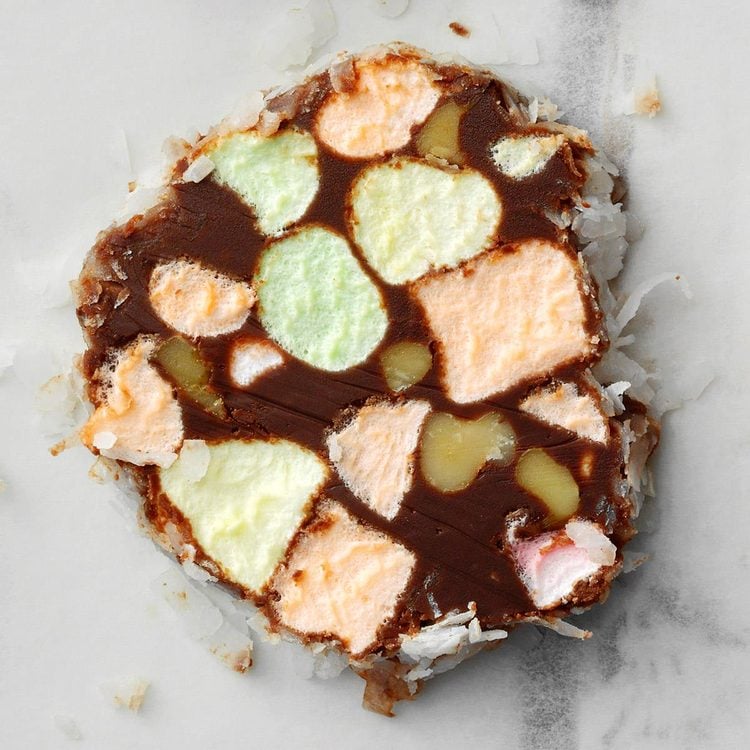 Church Window Cookies Recipe A close-up of a round slice of a multi-layered dessert with a dark chocolate base that holds irregular chunks of pastel-colored marshmallows in green, orange, yellow, and pink. These marshmallows are embedded unevenly inside the chocolate, creating a mosaic pattern with soft, fluffy textures. Around the edges, white shredded coconut pieces stick to the chocolate, adding a rough, flaky appearance. The slice rests on a white marbled surface, showing small crumbs scattered nearby. photo taken with an iphone --ar 4:5 --v 7