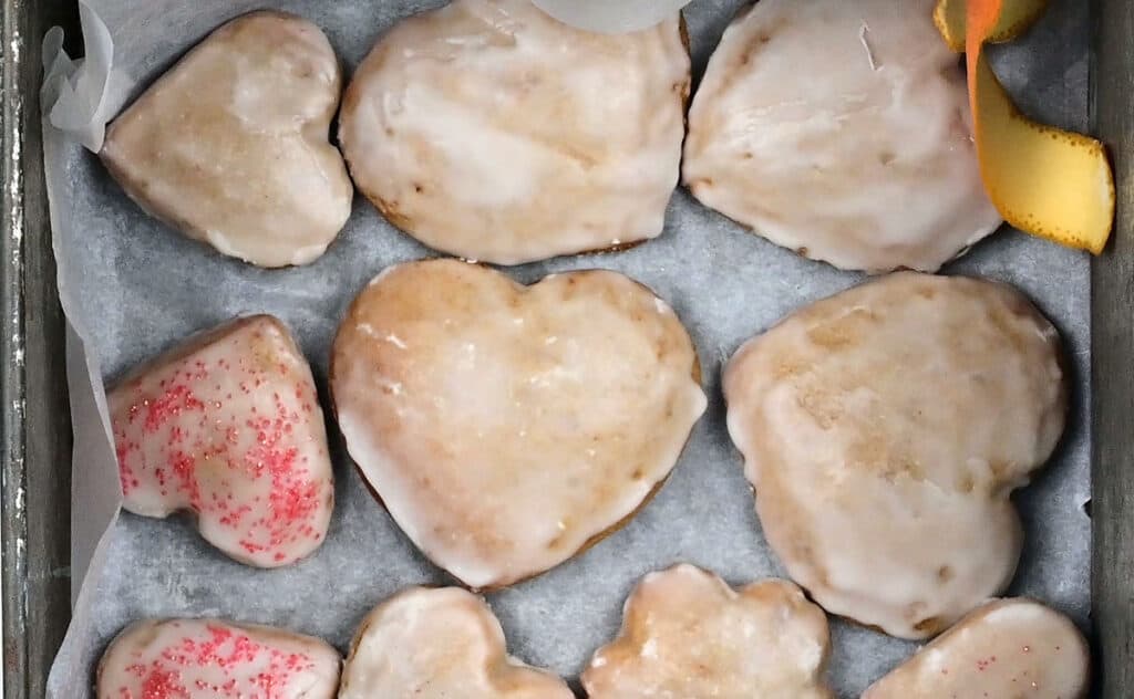 The image shows a baking tray lined with light gray parchment paper holding nine heart-shaped cookies. Each cookie is covered with a smooth, white icing layer that has a slightly shiny texture. Two cookies in the lower left corner have red sugar crystals sprinkled on top of the icing, adding a rough red texture. The cookies are arranged neatly with some touching each other. In the upper right corner of the tray, there is a curled orange peel, bright orange in color and textured. The entire scene rests on a white marbled surface. photo taken with an iphone --ar 2:3 --v 7
