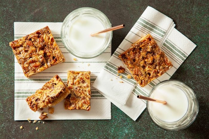Chocolate Toffee Pecan Bread Pudding Pie Recipe Four square nut bars with a golden brown top filled with chopped nuts and visible small pieces of nuts inside are arranged on white cloth napkins with green stripes. Two glasses of white milk with light brown straws stick out from each glass, placed beside the nut bars. One nut bar is broken into two pieces, showing a soft and slightly crumbly inside texture. All items are on a dark green surface, changed to a white marbled texture. photo taken with an iphone --ar 4:5 --v 7