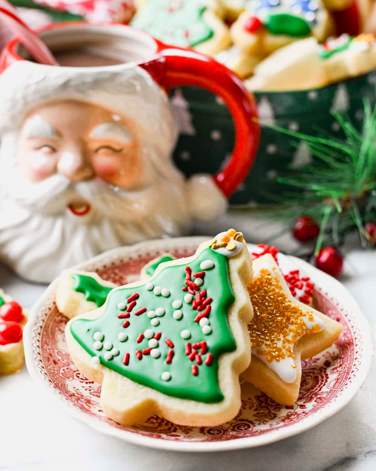 Soft Cutout Sugar Cookies with Edible Icing Recipe A white plate with red floral patterns holds two Christmas-shaped cookies in the foreground. The top cookie is a Christmas tree with smooth green icing covering its entire surface, decorated with small white round sprinkles and red elongated sprinkles scattered evenly, and small white and gold round details near the top edge. Underneath it, a star-shaped cookie is partially visible, covered with white icing and topped with shiny golden sugar crystals. In the background, there is a ceramic Santa face mug with a red handle filled with hot chocolate, and a white bowl filled with more decorated cookies on a white marbled surface with some green pine branches and red berries around. photo taken with an iphone --ar 4:5 --v 7 - Soft Cutout Sugar Cookies with Edible Icing, festive sugar cookies, easy sugar cookie decorating, tender holiday cookies, homemade cutout cookies