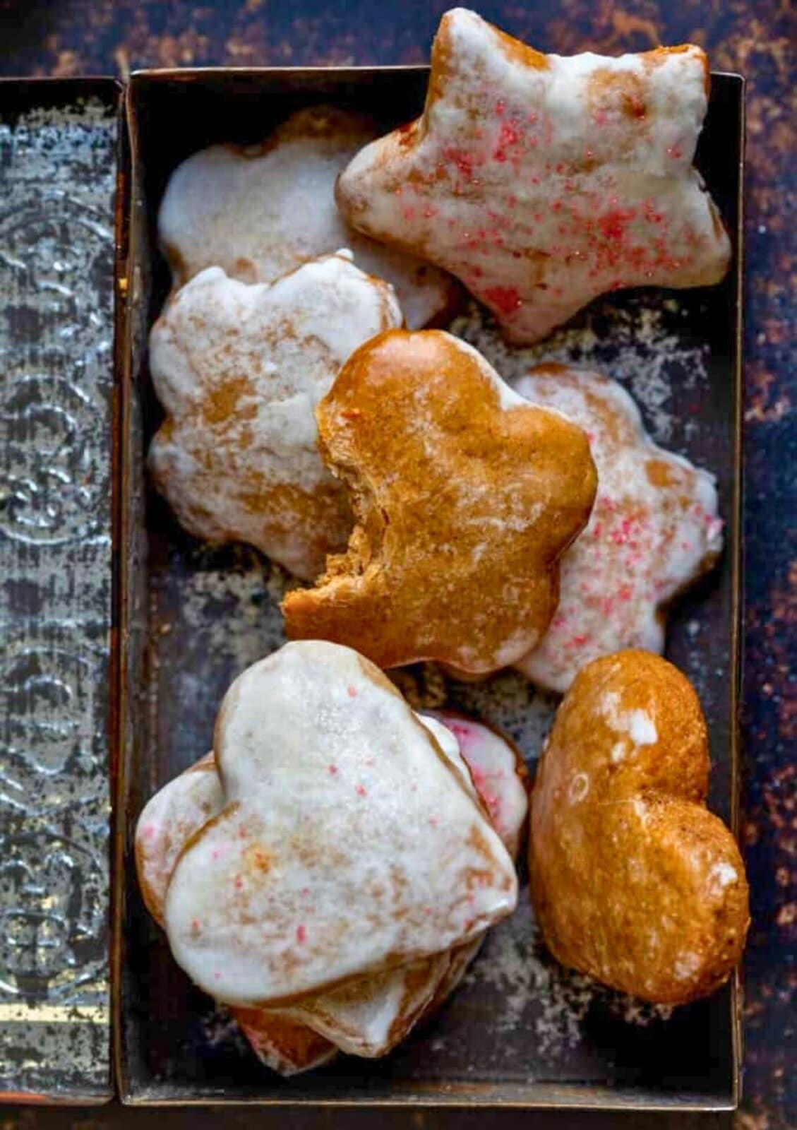 A metal rectangular tray holds several cookies with two main shapes: hearts and stars. Each cookie has a golden-brown base, with most covered in a thin white icing that has a slightly rough texture and some pinkish specks. One heart-shaped cookie is plain without icing, showing its smooth, golden top with a bite taken out of it. The faces of the cookies are slightly shiny, and the edges are rounded. The tray sits on a white marbled surface. photo taken with an iphone --ar 2:3 --v 7