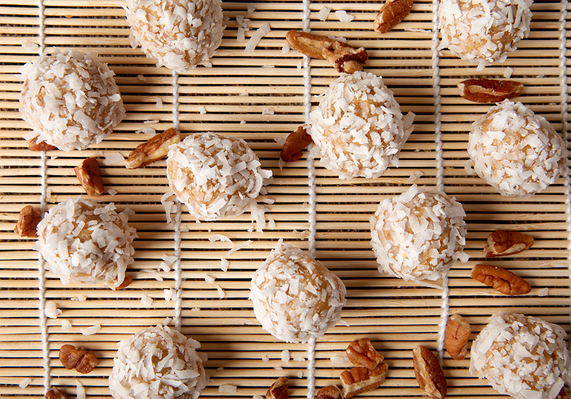 The image shows several round energy balls coated with white shredded coconut, placed neatly on a bamboo mat with thin white strings running vertically underneath them. The energy balls have a light beige color visible under the coconut flakes, giving a rough texture on the surface. Scattered around and between the balls are pecan halves with their warm brown and slightly glossy look. The whole setup is on a white marbled surface, giving a clean and bright background. photo taken with an iphone --ar 4:5 --v 7